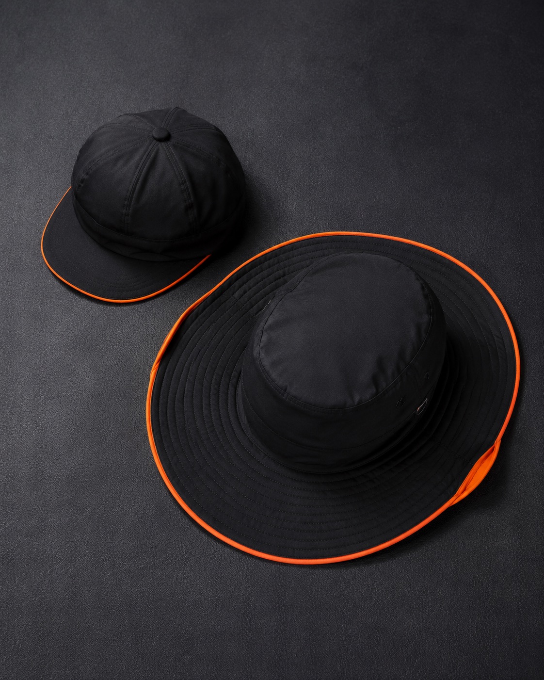 Cricket baggy cap beside a wide-brim sun hat, flat lay on dark charcoal