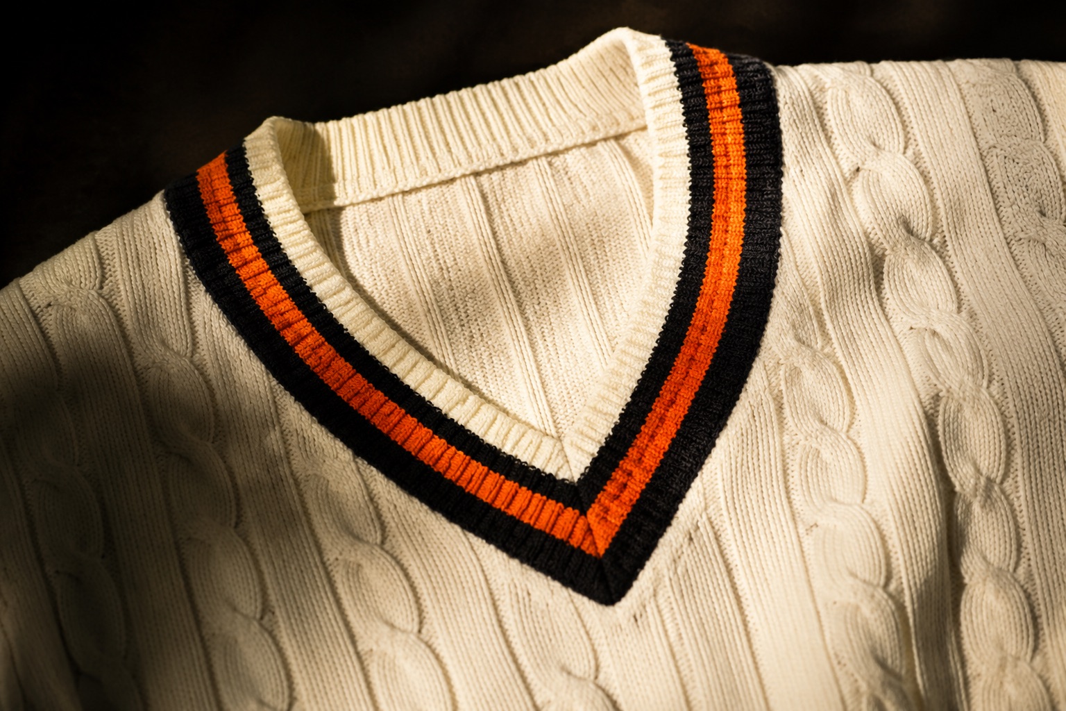 Close detail of a cream cricket sweater showing cable knit and coloured tipping at the V-neck